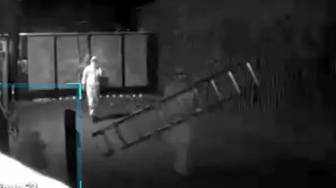 CPS A grainy black and white still from CCTV. There are two people in white forensic-style suits inside a building. The man in the foreground is holding a ladder.
