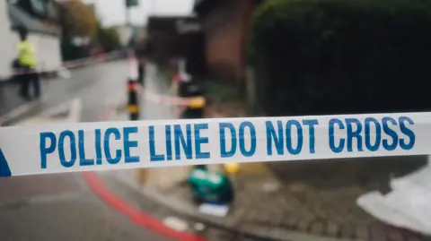 A photo of the white and blue police cordon tape and a street in the background, in the place of the crime scene.