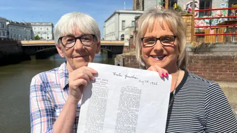 An image of two older women holding a newspaper clipping from 1926. The woman on the left has short hair and black glasses. She is wearing a blue and pink shirt. The woman on the right is wearing a stripy black and white top and wearing black glasses.