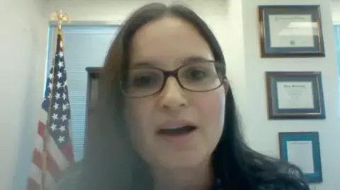 Reuters Aileen Cannon, who has long black hair and is wearing glasses, answers questions via Zoom during her Senate nomination hearing in 2020, with an American flag on a pole over her right shoulder and three framed certificates over her left shoulder