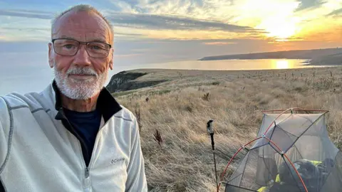 Graham Eaves A man with white hair looking at the camera with a white jumper. He has glasses and is on the cliff edge with a tent and walking pole with the sun setting behind him.