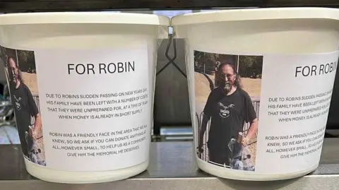 Kathryn Stonehouse Two white buckets placed on a stainless steel counter, with a photo of Robin Stonehouse and a message from his family about the fundraising.