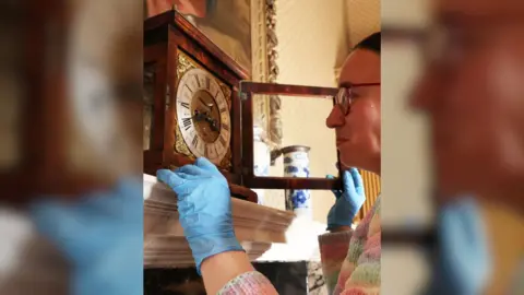 National Trust Images/Janine Sterland Picture of a brown haired woman with glasses looking at a clock on the top of a fireplace. 
