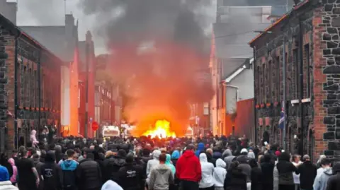 Pacemaker is a group of people who stand on the street with broken houses. There is a police boat on fire in the background.