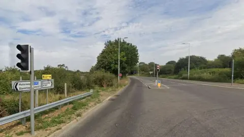 Google The junction of Graveley Road/High Street and North Road/B197. There are white road signs on the left pointing in opposite directions, one says Motorway A1(M) Stevenage and the other says Graveley B197. There is a crash barrier below the signs and traffic lights next to it, as well as traffic lights in the middle of te photo. Trees line the road alongside street lamps.