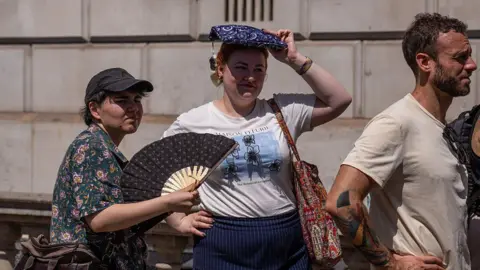 A woman fans herself as another shelters from the sun as they wait to take a picture in front of a phone box in Parliament Square. In front of them is a man wearing a beige top.