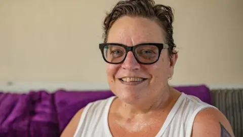 East Anglian Air Ambulance Ruth Barron, with short hair swept back, and wearing glasses, a white vest top, sitting down on a sofa with a purple blanket in the background.