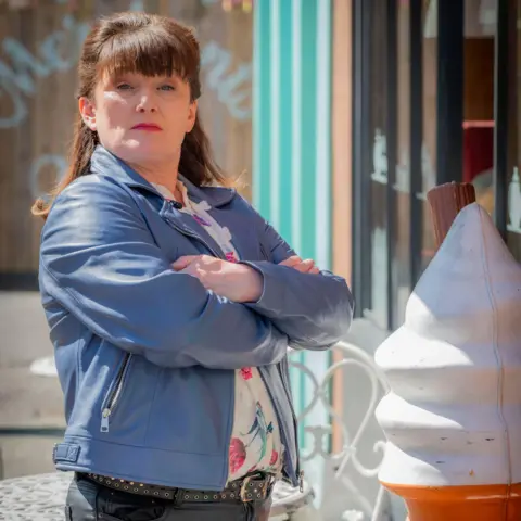 BBC Studios Drama Scarlett O'Hara person stands with arms folded beside a large decorative ice‑cream cone outside a storefront.
She wears a blue jacket and dark trousers, positioned near white metal café furniture on a pavement.
Behind the person, a striped shopfront and glass window suggest a street‑side dessert or retail setting.