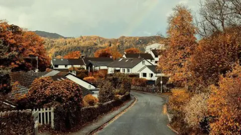 BBC Weather Watchers/Keri A shot looking down a small residential street where white houses with pitched-roof houses are surrounded by autumnal trees and shrubs, with the fells visible in the distance. 