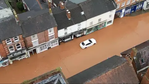 Darren Thompson An overhead shot looking down on a street flooded with muddy water. A white car sits in the water. The street is lined with shops and it is clear that the water is very high.