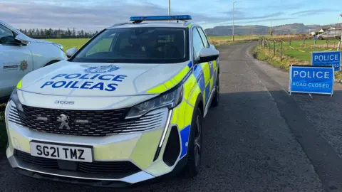 A police car sits beside two signs reading Police Road Closed. Fields are visible in the background