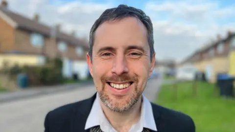 Simon Dedman/BBC Chris Vince standing in a residential street. He has short black hair, a short beard and is smiling while wearing a navy blazer and light grey shirt.