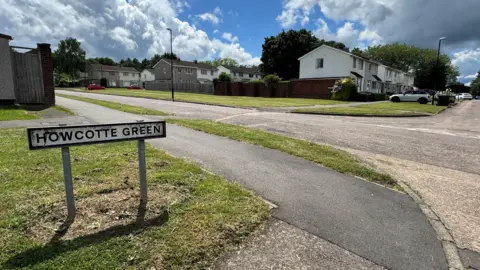 A residential street with a “Howcotte Green” sign, houses and a path under a partly cloudy sky.
