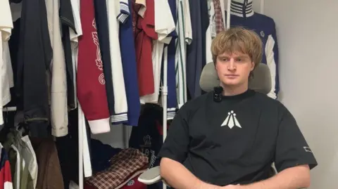 Alexei Hamblin has blond shaggy hair. He is looking off to the side and is sitting in front of a rail of sportswear.