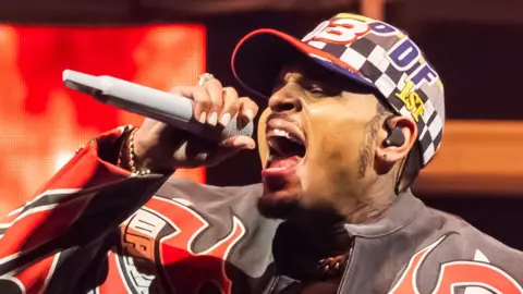 Chris Brown in a red, white and black patterned leather jacket and patterned baseball cap, singing into a microphone on stage at the Tycoon Music Festival at Little Caesars Arena on April 19, 2025 in Detroit, Michigan.
