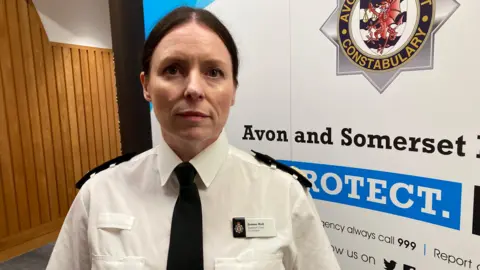 Assistant Chief Constable Joanne Hall pictured wearing her uniform of a white shirt with two black shoulder patches, a black tie and a name badge. She has dark hair tied back in a low bun or ponytail and is looking directly at the camera. There is a large sign with the Avon and Somerset Police logo directly behind her. 