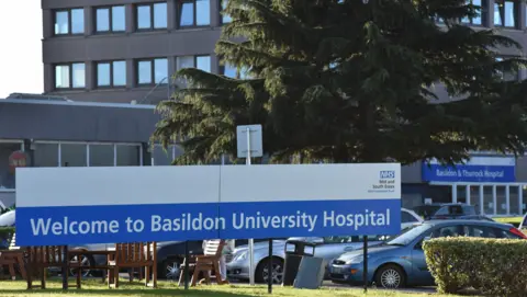 The exterior of Basildon Hospital, where there is a large blue and white sign advertising its name. Behind it are wooden chairs, a tree and the main hospital building, which is dark grey.