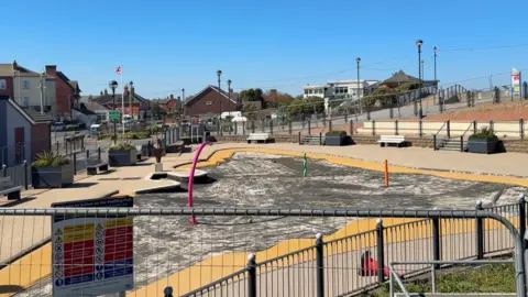 View over metal fencing at a children's paddling pool which has had its surface ripped up and has no water in it