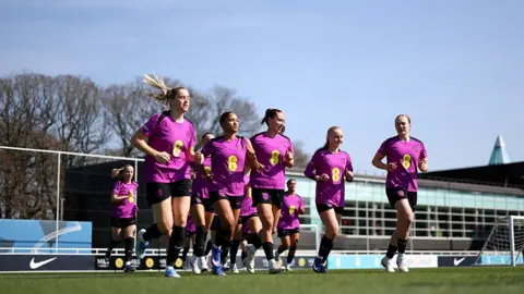 Football Association England Women wear bright purple football tops with yellow EE branding. They are seen running on a football pitch. 