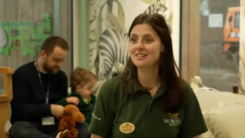 DJ McLaren/BBC Natasha pictured inside the sensory room, with a child and adult behind her sitting and playing with a soft toy. She looks animated and wears a forest green Woburn T-shirt, with a tiger as its logo.