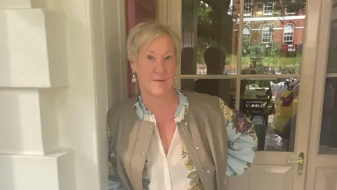 Deborah Clark Deborah Clark in a cream and floral-patterned blouse and an olive collarless gilet standing in the entrance to Southernhay House and Hotel