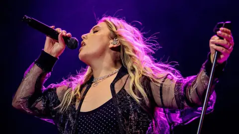 Artist Ella Henderson performing at a concert. She is wearing a black outfit, part of which appears purple from the stage lighting.