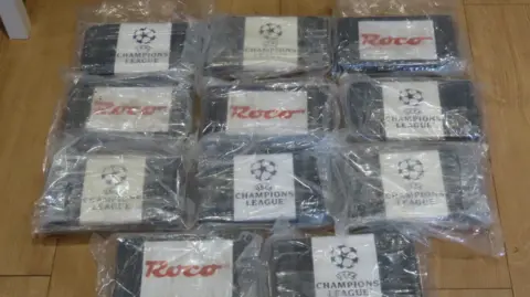 National Crime Agency A selection of drugs wrapped in plastic with labels saying 'Champions League' and Roco. They are arranged on a wooden floor.