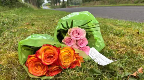 Floral tributes left on a roadside, near the scene of a fatal gun attack in Maguiresbridge.  A bunch of orange roses and a bunch of pink roses lie on the grass. A handwritten note is attached. 