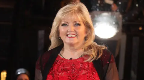 PA Media Linda Nolan had long blonde hair and was wearing a red dress with a black cardigan.
