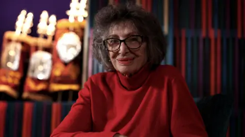 Dorit Oliver-Wolff looks at the camera and is wearing a red roll-neck jumper. She has glasses on and bobbed dark grey hair.