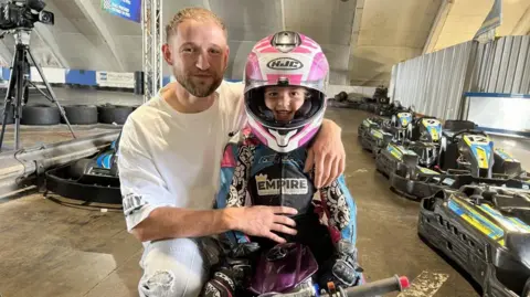 A young man in a white t-shirt crouching down next to a little girl on a mini-moto in a purple racing suit and helmet. 