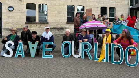 Campaigners in favour of the lido's reopening pose ahead of the Stroud District Council meeting. They kneel behind large letters which spell out "SAVE OUR LIDO". 