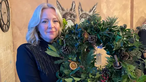 BBC/Amelia Riley Serena Coope - a woman with shoulder-length blonde hair and a black sparkly jumper on stands looking into the camera. She is holding a completed Christmas wreath, decorated with orange slices, pinecones, and cinnamon sticks.