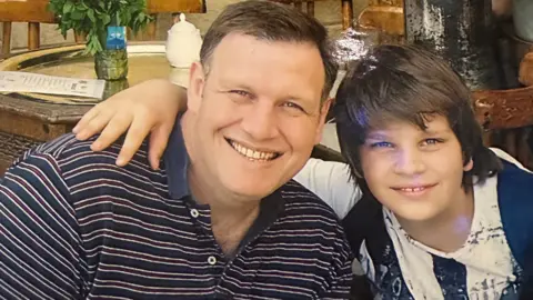 Conor Buckett, pictured as a young boy has his arm around his father, who is wearing a striped polo shirt. Both of them are looking into the camera. 