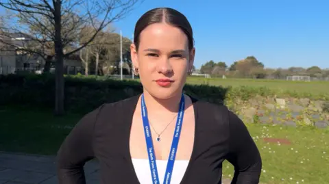 A teenage girl with a slick back brunette pony tail. She is wearing a white top with a black cardigan. She has a blue student lanyard on and big hoop earrings on 