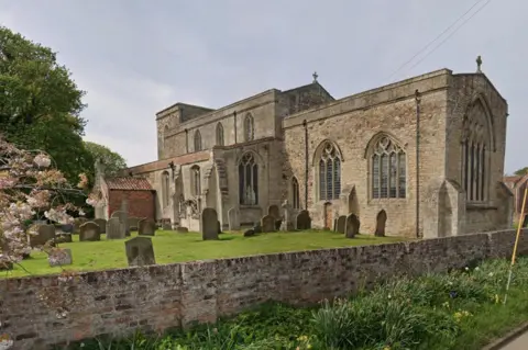 Google An ancient, brick-built church with large arched windows and a squat tower, standing in green grounds with gravestones around it. In the foreground is a low brick wall.