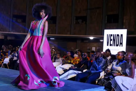 KB Mpofu/Reuters A model in a long pink dress with a strip big collar on the catwalk being photographed by members of the audience at the I Wear My Culture fashion show in Bulawayo, Zimbabwe - Saturday 30 August 2025.
