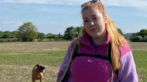 A woman with long strawberry blond hair is wearing a bright pink and purple fleece. She is stood in a field with a black and tan coloured dog in the background