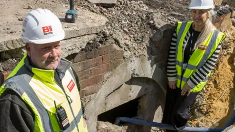 Barron Media Jason Harrison and Lauren Amerigo are standing either side of a cracked open entrance into an underground air raid shelter. Both are wearing white helmets and yellow high-vis vests.