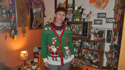 BBC Lisa Watts, a smiling woman wearing a green Christmas cardigan with Santa and snowman designs stands in a vintage-style living room filled with holiday decorations, figurines, tinsel, and a small decorated tree on a shelf behind her.