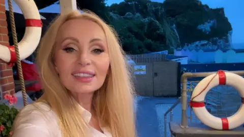 Supplied Ewa Larsson smiling in a selfie. A coastal scene in fading light is visible behind her.