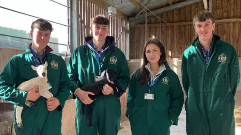 BBC A group of farming students in a barn they are all wearing green overalls and two of them are holding lambs, one black and one white. the students consist of three male and one female.