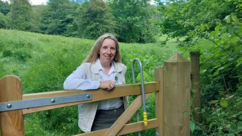 Deborah Fox A woman with blonde hair leans against a wooden gate in the middle of a field. She is wearing a white shirt and a beige jacket over the top.