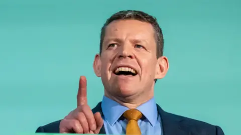 Rhun ap Iorwerth, a man with short brown hair in a navy suit, speaks while standing at a lectern.