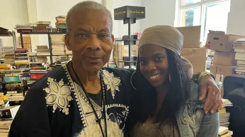 An elderly man stands in a bookshop with his arm around a young woman in a bookshop, surrounded by piles of books. 