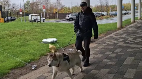 Insp Andy Robinson in a waterproof black coat, police cap and black gloves holding a lead to attached to a white husky dog. 