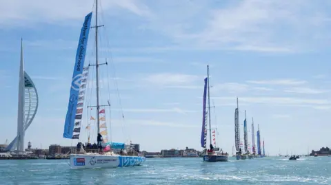 Supplied Sailing boats sail into Portsmouth in a line. The sun is out. The water is saphire blue. 