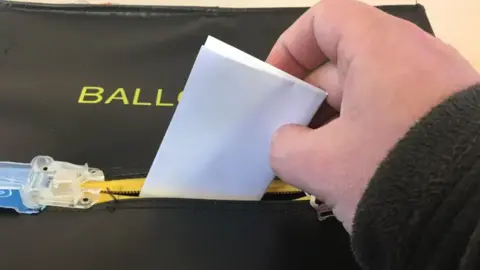 BBC A hand placing a folded piece of paper into a black ballot box.
