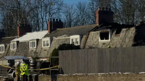 Houses with blackened areas taped off with yellow tape. Firefighters stand by the cordon and it is a bright sunny day with blue skies 