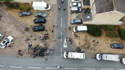 John Fairhall/BBC. Una fotografía aérea muestra a varias personas reunidas en una esquina, junto con otras en otros puntos de la calle. Se observan numerosos vehículos estacionados en la calle y en entradas de garajes.